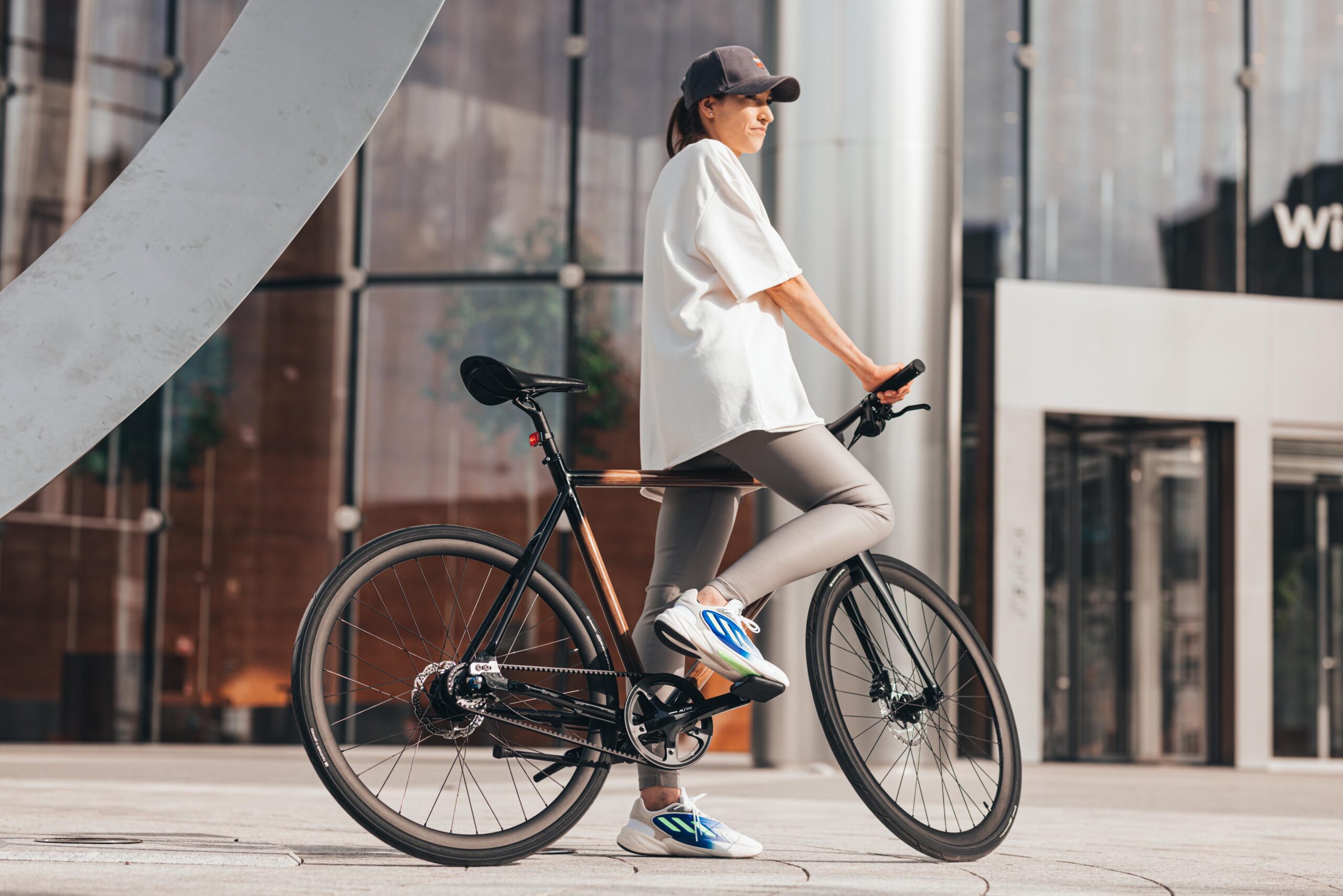 Sporty dressed girl in the middle of a square surrounded by glass walls standing with bicycle with foot on the pedal looking straight ahead. The sun illuminates them. Bicycle with a classic male frame, covered with natural wood.
